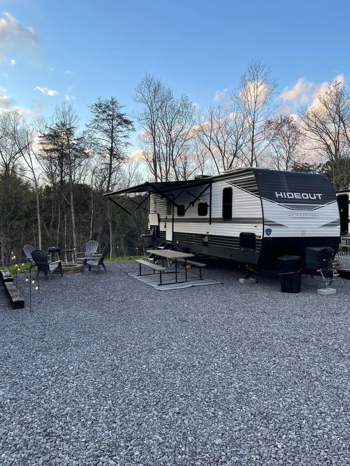 The image showcases a modern Keystone Hideout RV set on a gravel pad, surrounded by a cozy outdoor seating area and a glowing fire pit. The setting is tranquil, with the soft light of sunset casting a warm glow over the space, inviting guests to gather for stories, s'mores, and stargazing against a scenic Smoky Mountains backdrop.