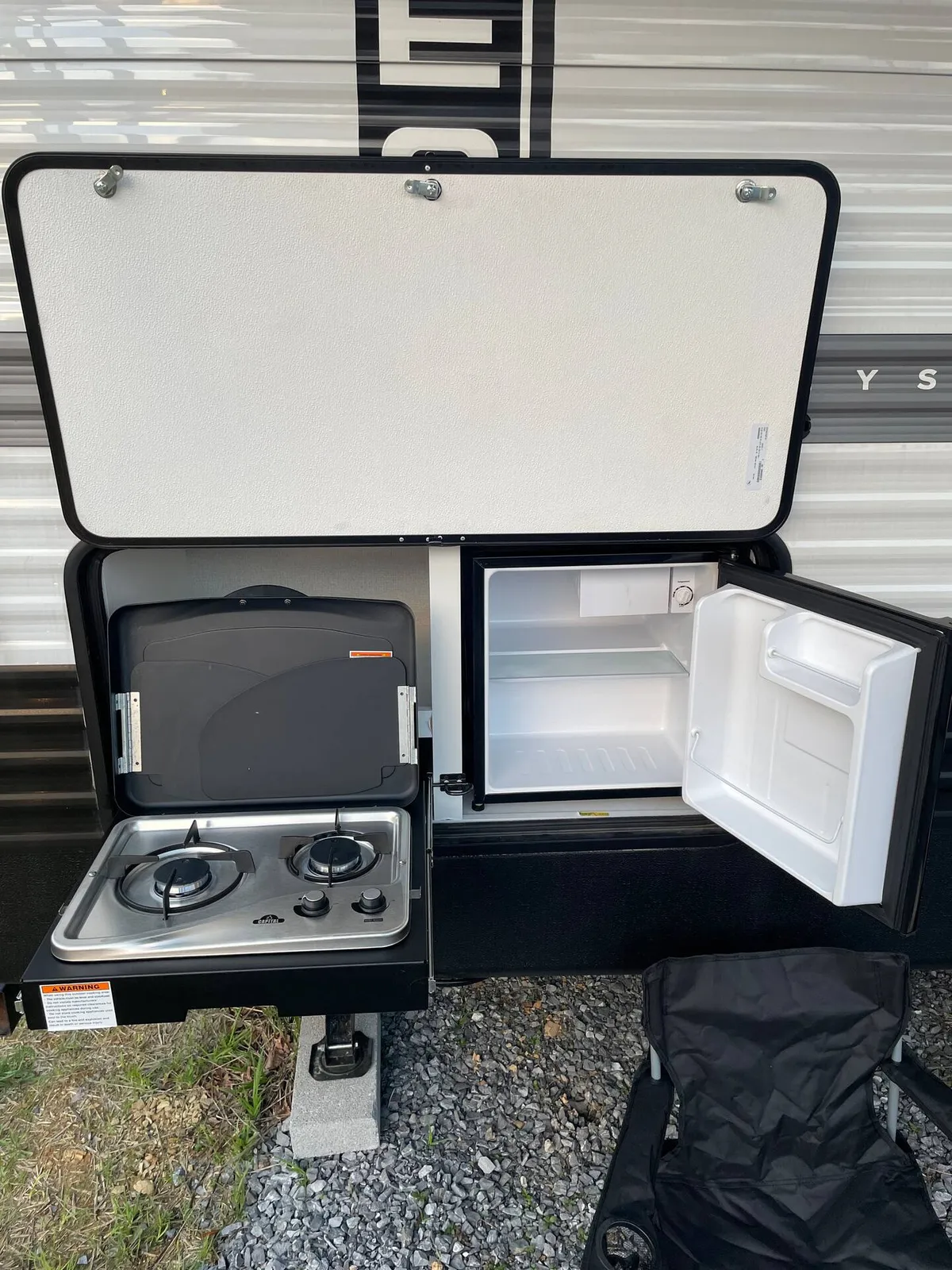 This image shows an outdoor camping setup featuring an outdoor cooking stove and compact refrigerator situated on a gravel pad. A portable chair sits nearby, inviting relaxation. The scene captures the spirit of cooking outdoors, with hints of early morning light and the possibility for shared meals or snacks by a fire.