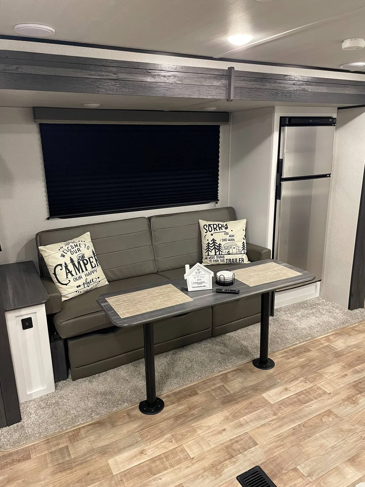 A cozy dining nook with a table, sofa seating, and decorative pillows, set beside a visible refrigerator. The space is inviting, ideal for sharing meals, relaxing, or planning family adventures. The refrigerator subtly anchors the kitchenette's presence in the room.