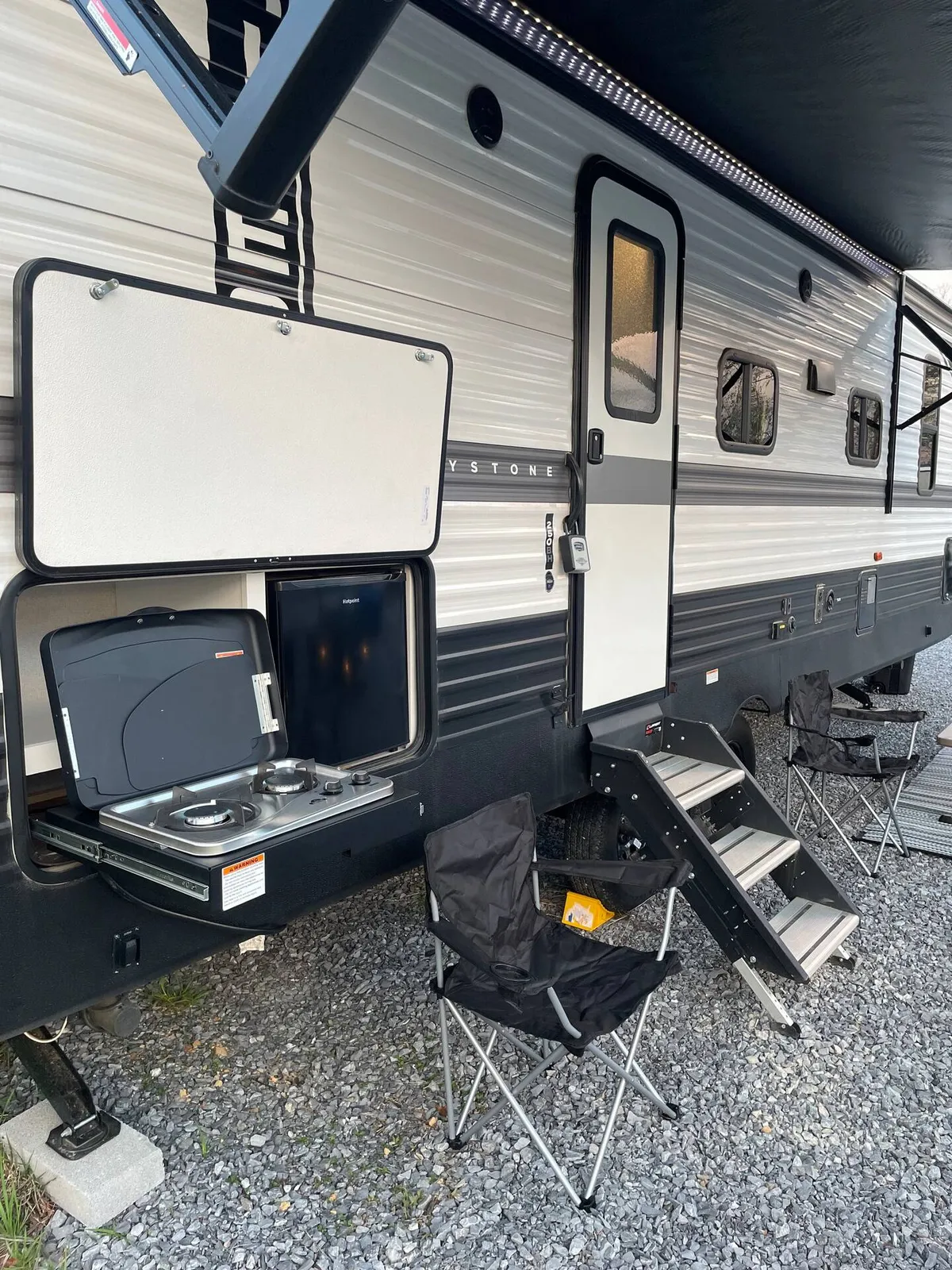 Here, the exterior of a camper is paired with an outdoor cooking area and several folding chairs set up nearby. The scene suggests a welcoming social atmosphere, perfect for gathering, chatting, and enjoying meals or evening stargazing under the open sky.